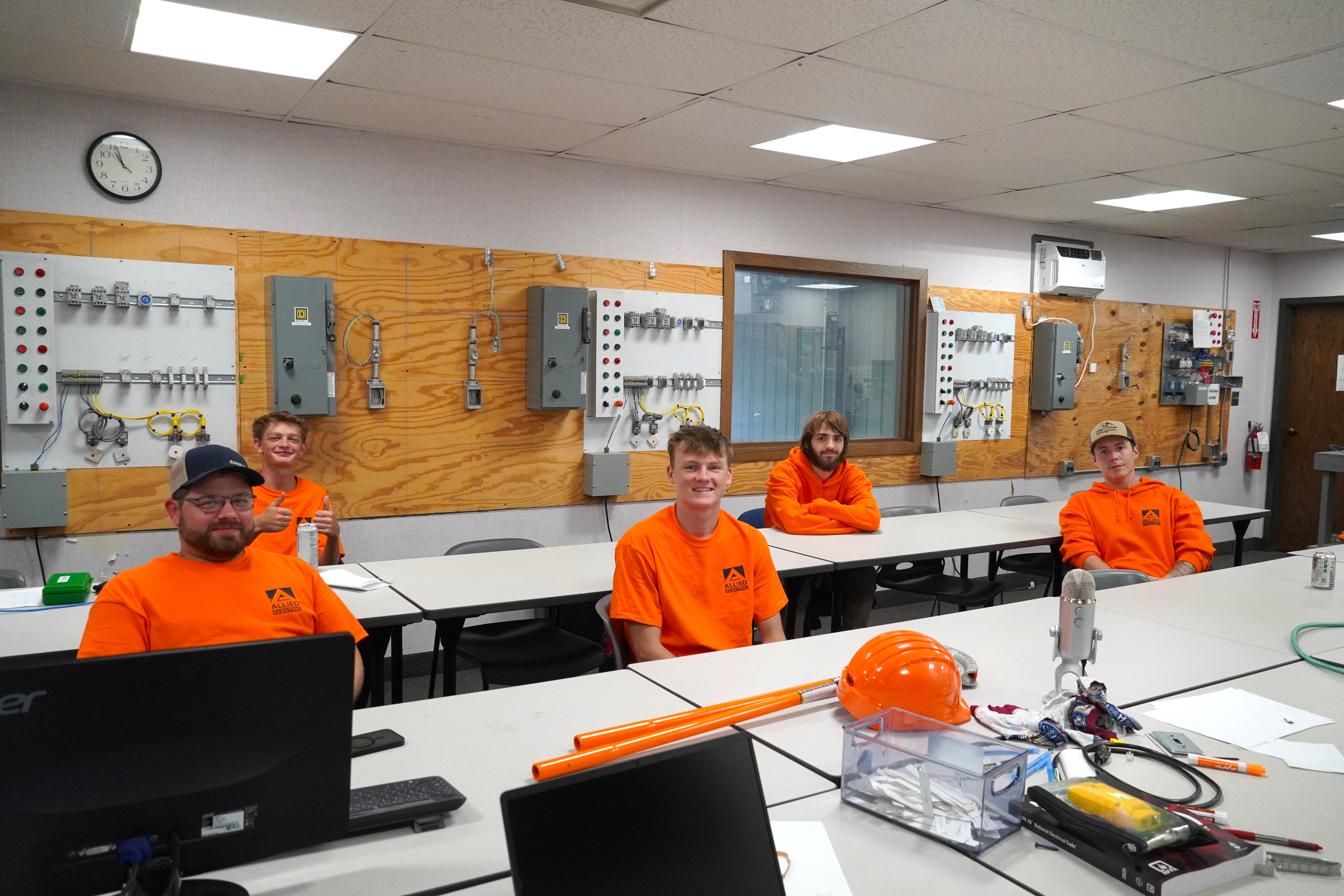 Allied Electric apprentices in orange safety shirts sitting in electrical training classroom with hands-on training panels