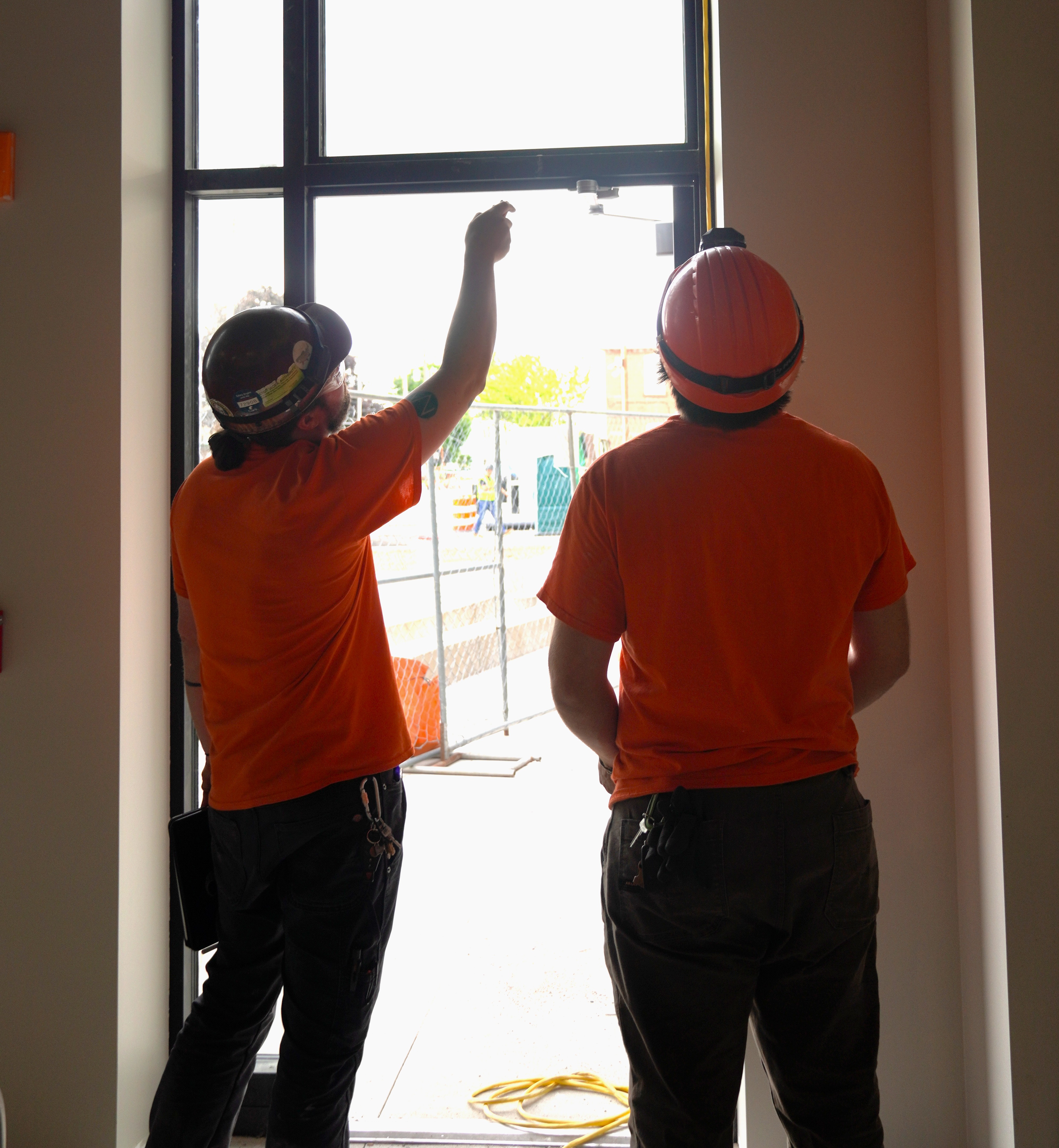 Two Allied Electric workers in orange safety shirts and hard hats collaborating at a construction site window