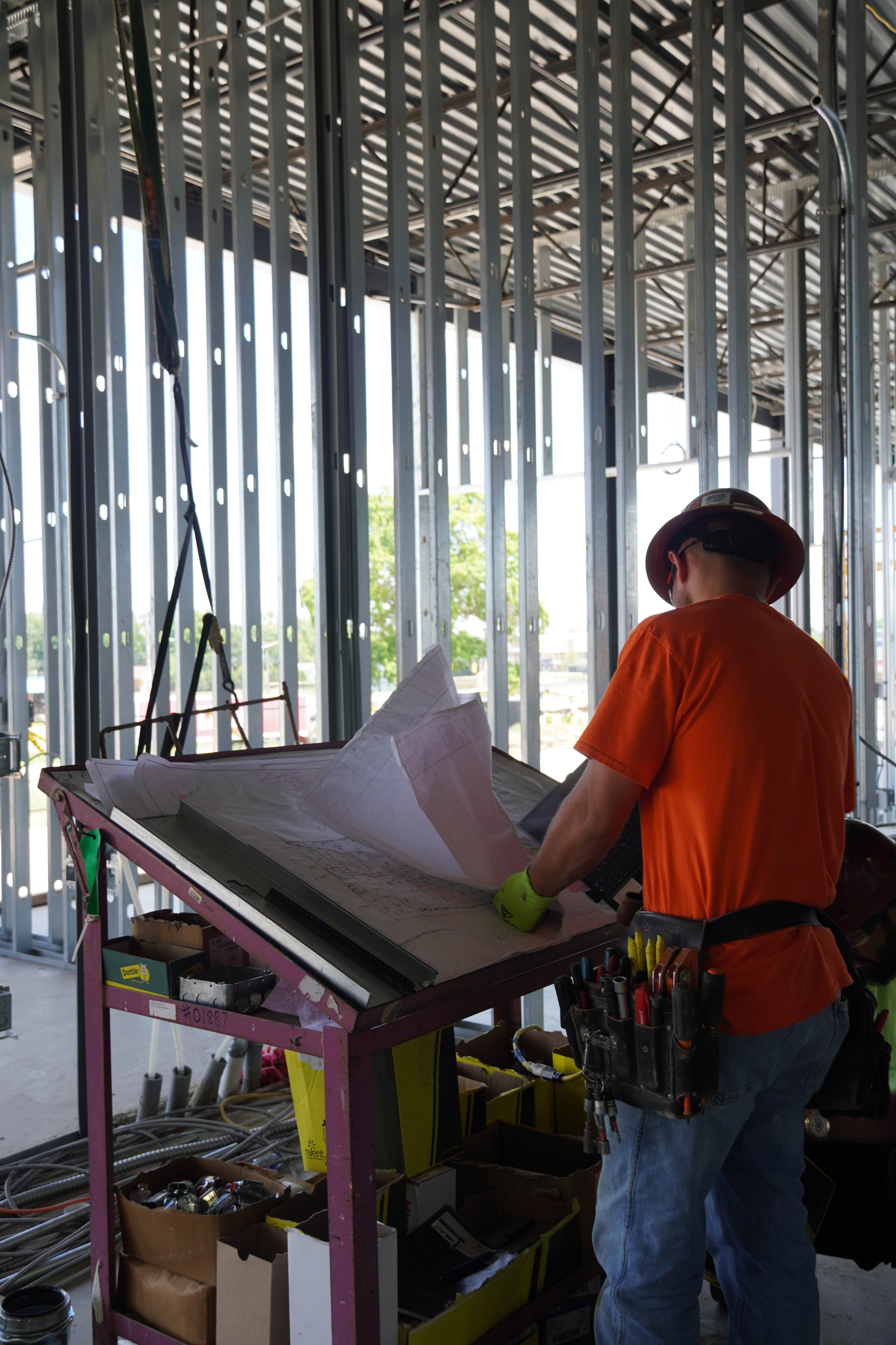 Allied Electric worker in orange safety shirt and hard hat reviewing construction blueprints on drafting table at construction site